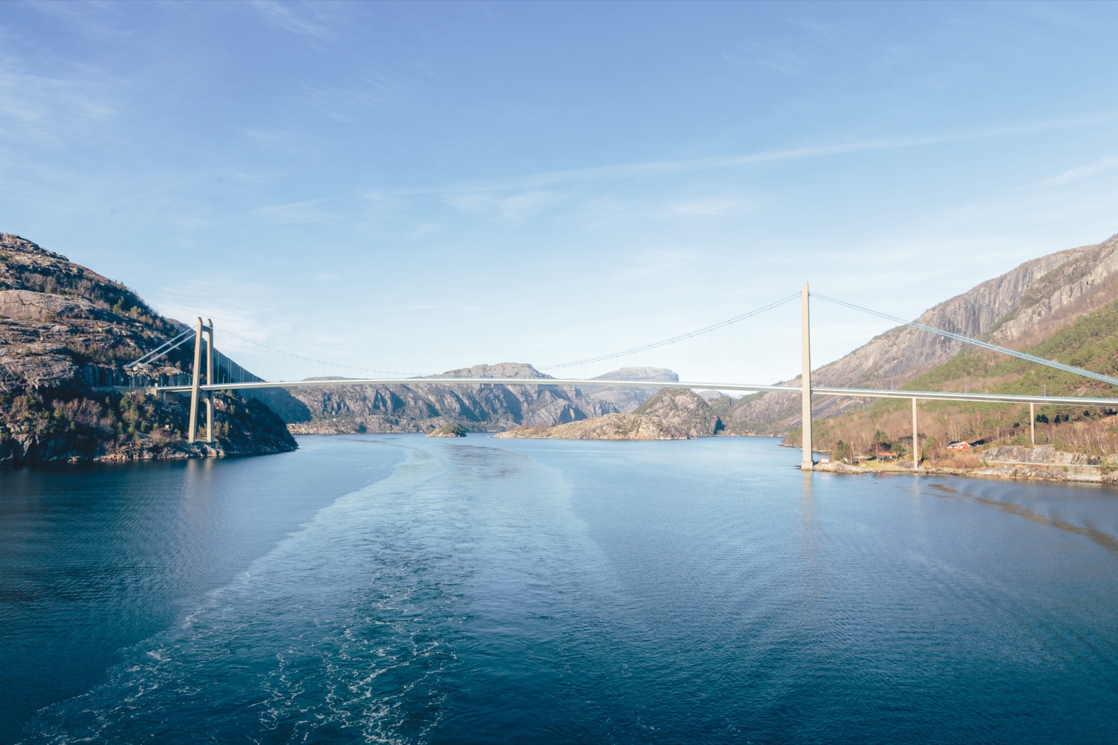 En hengebru over en fjord mellom steinete åssider, sett fra kjølvannet til en båt