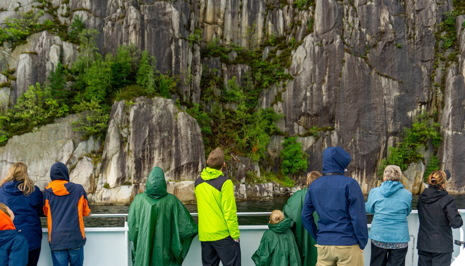 Passasjerer i regnjakker ved rekka på en båt, som ser opp mot en loddrett fjellvegg